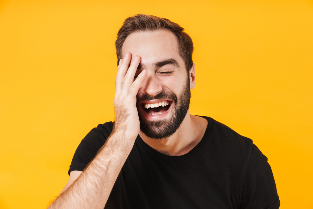 man laughing, covering one side of his face with his hand, wearing a black shirt, on yellow background