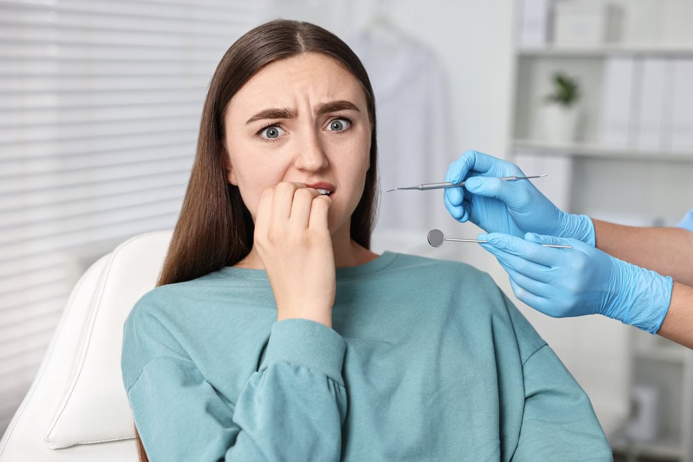 Patient sits in a dental chair looking anxious as blue-gloved hands present dental instruments near her mouth for an examination or procedure.